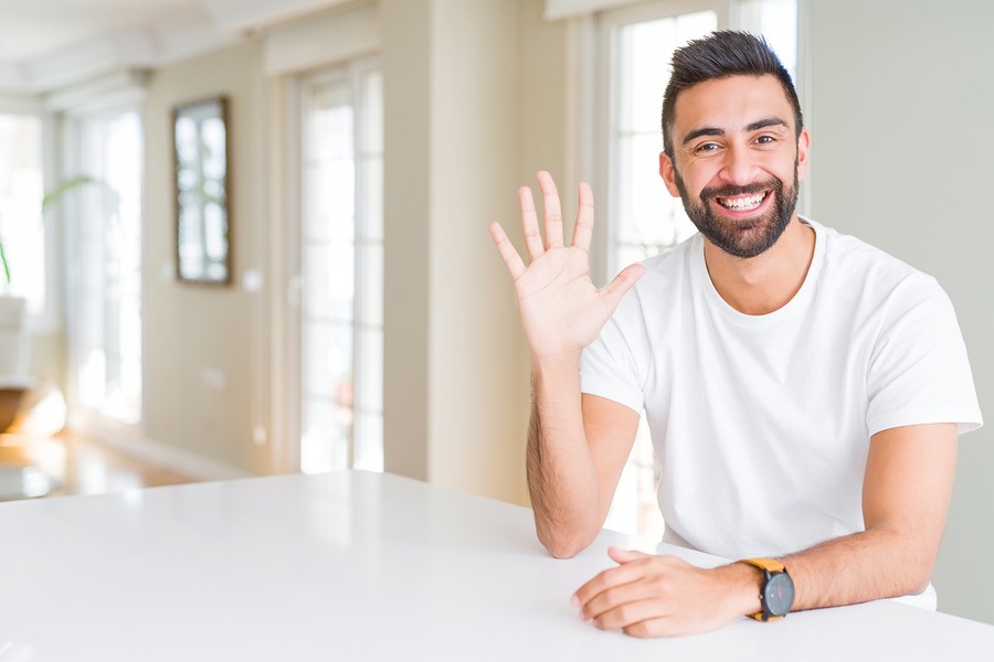Handsome hispanic man casual white t-shirt at home showing and pointing up with fingers number five while smiling confident and happy.