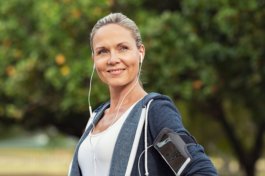 Portrait of a smiling healthy woman with earphones standing in park after running. Proud mature woman looking away after work out. Portrait of satisfied mid woman after fitness exercises.